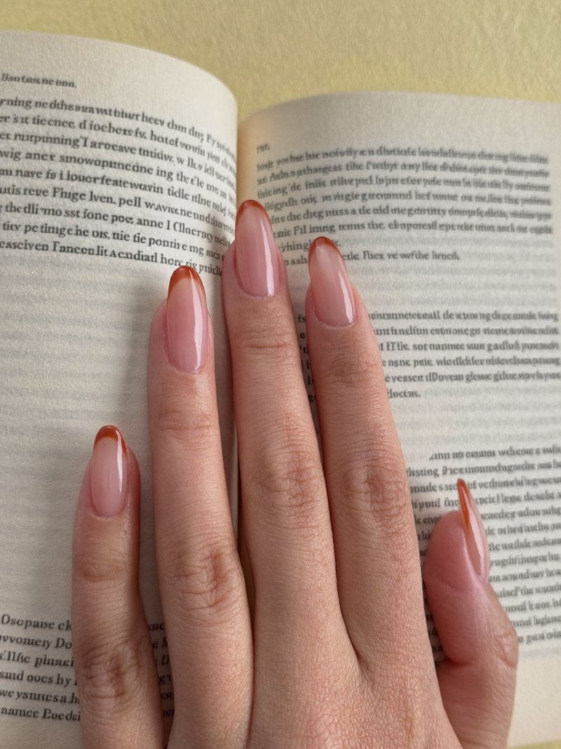 Short almond nails with a sheer base and thin burnt orange French tips