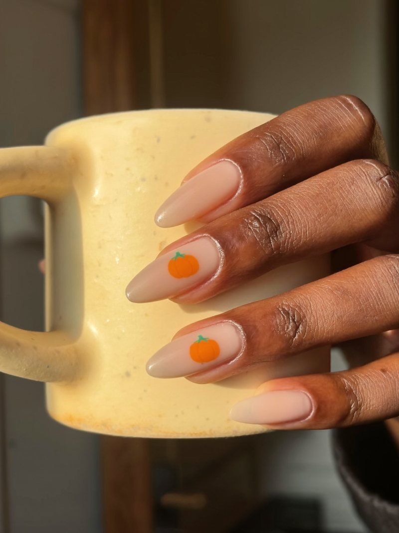 Short almond nude nails with a small orange pumpkin on the ring finger